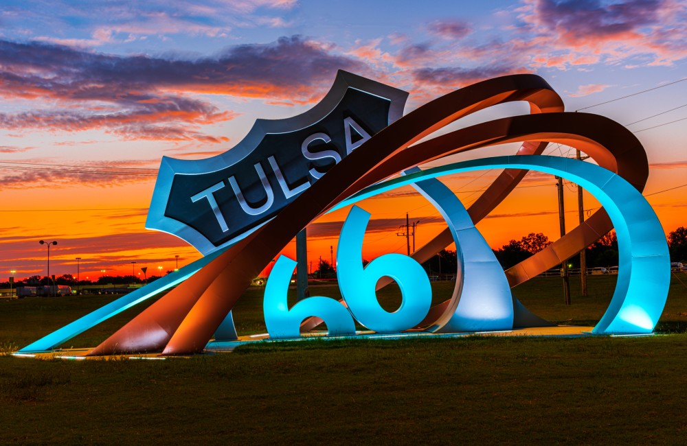 Large sculpture consisting of three metal bands rising from the ground (left) and curving around a replica of a "Tulsa Route 66" sign (center). The sculpture is located in an open grassy area and is illuminated by ground lights.