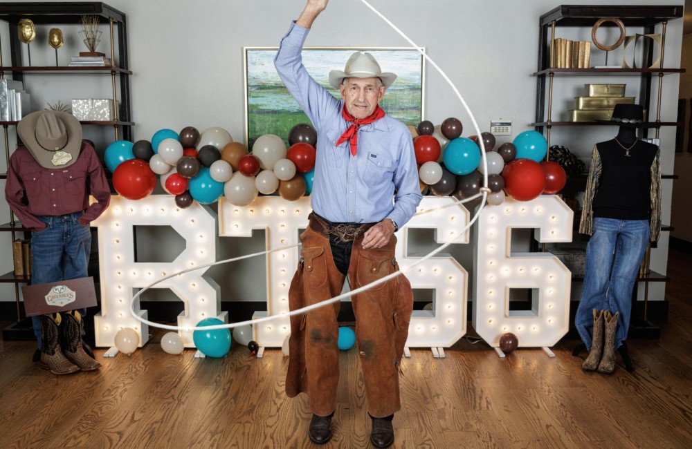 An older man wearing traditional cowboy clothing twirls a lasso while standing in front of illuminated letters spelling "RT66."