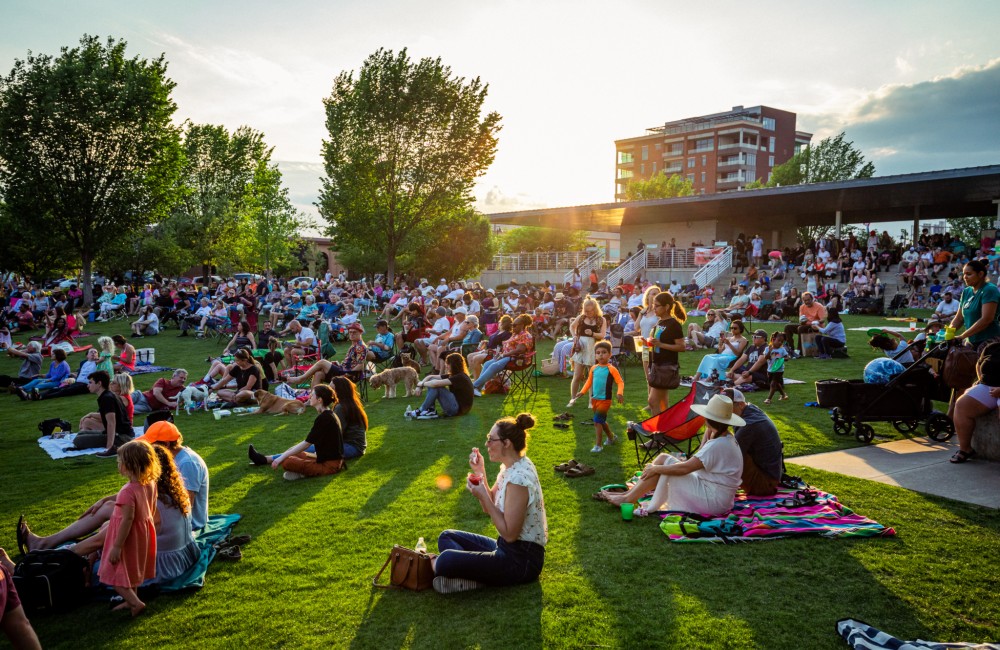 People sit on a grassy lawn; trees, a tall building, and light from the setting sun are visible in the background.