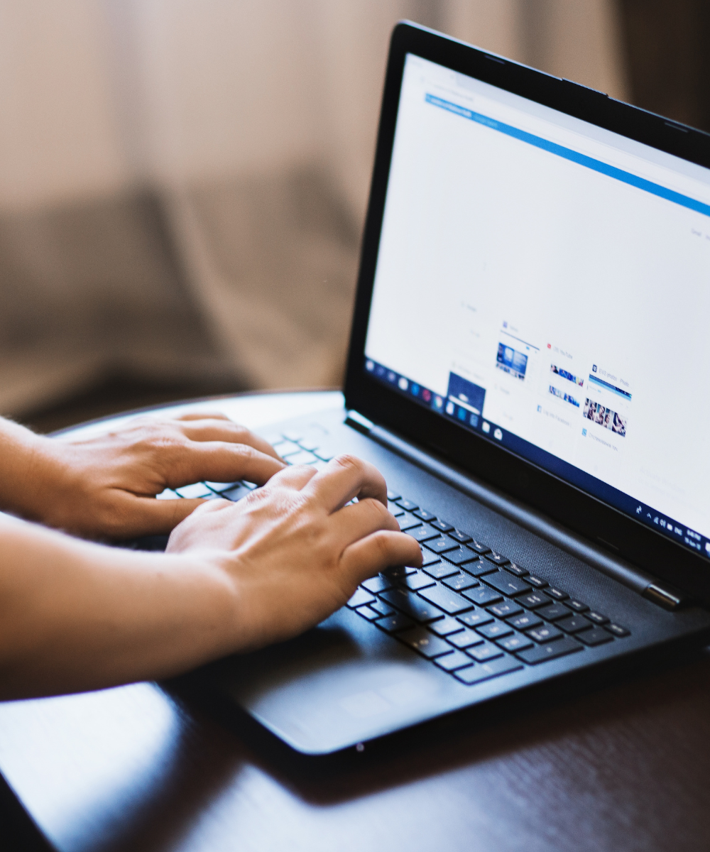 Hands typing on a laptop keyboard showing Facebook feed on laptop screen