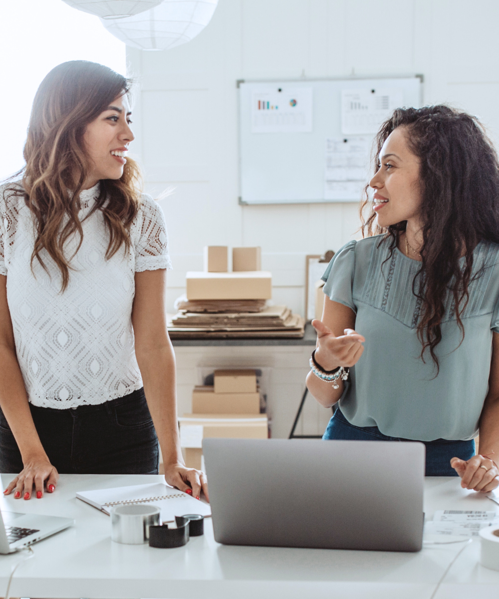 Stock image of two women small business owners in store having a conversation at laptop computers on desk table workstation