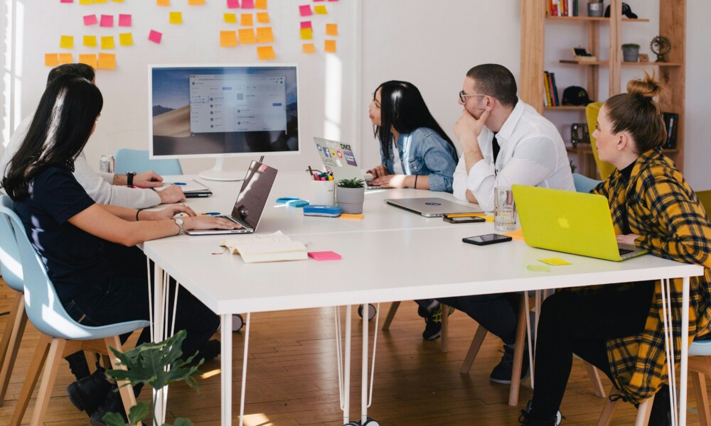 People sitting around a table with laptops looking at a board with sticky notes