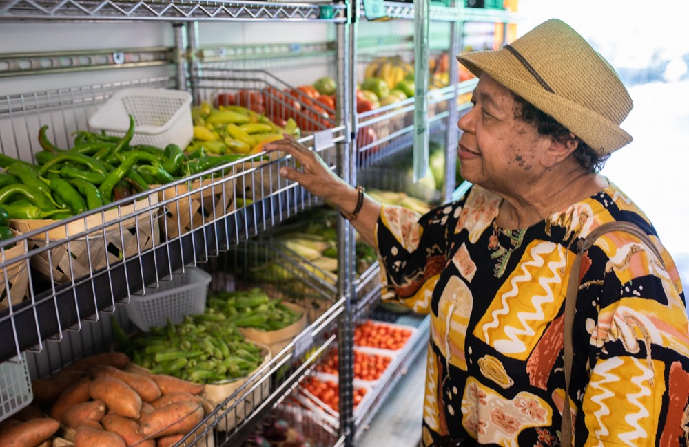An older woman reaches for green peppers while shopping at a mobile farmers market whose shelves are filled with fresh produce.