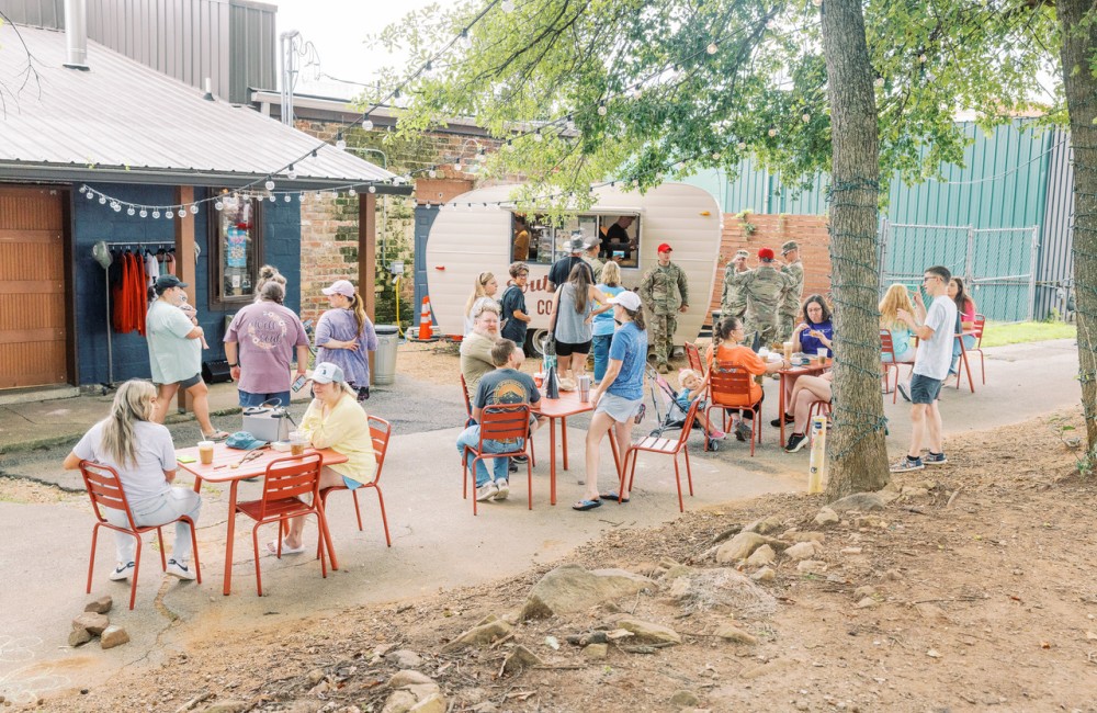 In an outdoor public seating area, people line up to place orders at a coffee shop operating from a small vintage-style travel trailer, while others enjoy their beverages while sitting at tables.