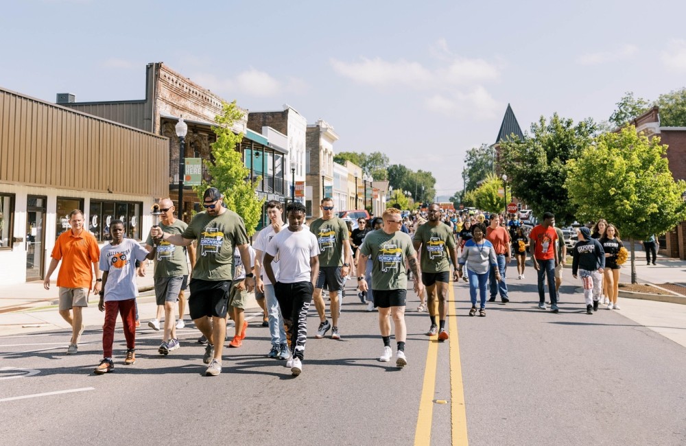 A large group of people of all ages walk down the center of a downtown road, toward the viewer.