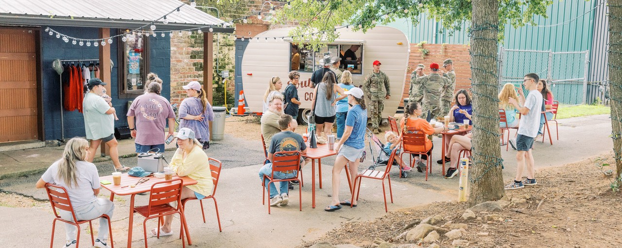 In an outdoor public seating area, people line up to place orders at a coffee shop operating from a small vintage-style travel trailer, while others enjoy their beverages while sitting at tables.