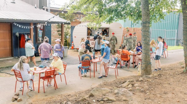 In an outdoor public seating area, people line up to place orders at a coffee shop operating from a small vintage-style travel trailer, while others enjoy their beverages while sitting at tables.