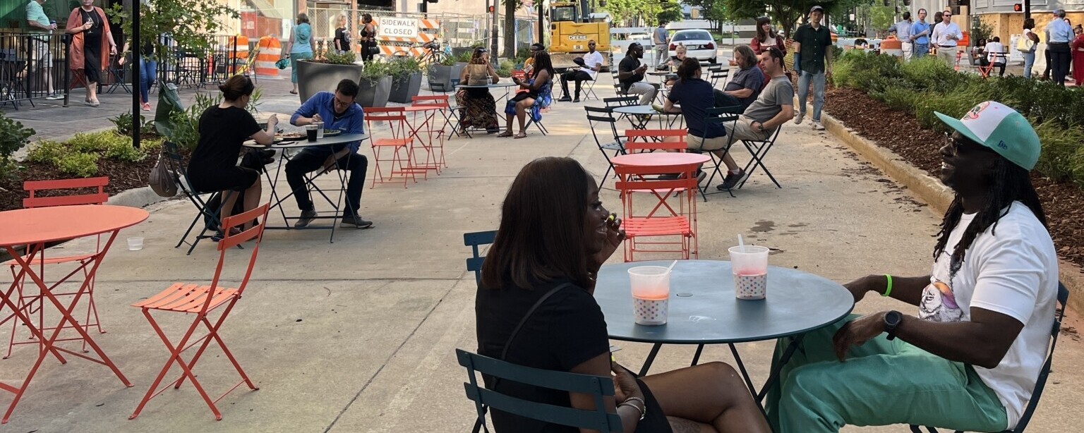 People sitting at café tables set-up along a street lined with tall office buildings.