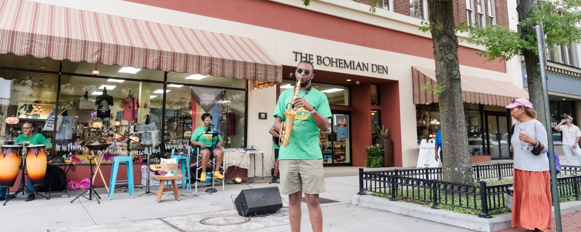 A saxophonist, drummer, and vocalist set-up on a wide sidewalk perform for pedestrians.