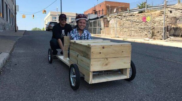 A woman wearing a helmet sits in a wooden derby car, a man behind her gets ready to push the car.
