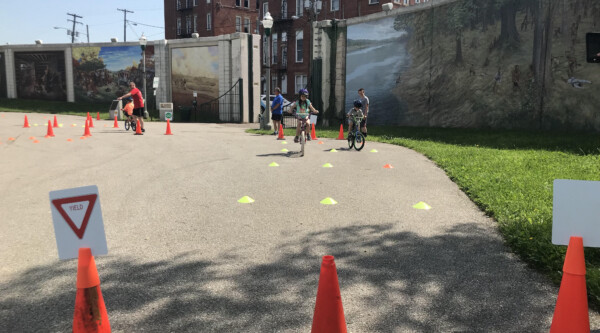 Orange pylons guide the way for young bicyclists on a sidewalk.