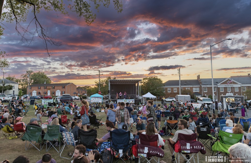 Large group of people sit on grassy lawn listening to music on outdoor stage at sunset.
