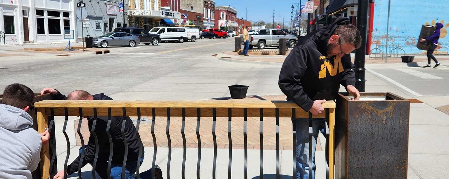 Three people working together to install a planter and fence, while in the background another person carries a square planter and main street lined with historic buildings are visible.