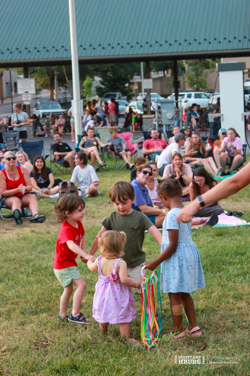Group of four young children hold hands in circle on lawn where group of people gather sitting on lawn chairs and blankets.