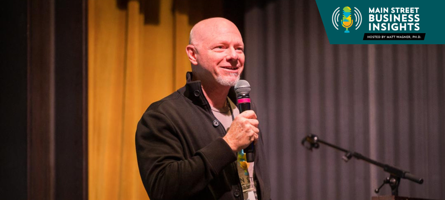 Man standing in front of a podium on a stage holds a microphone.