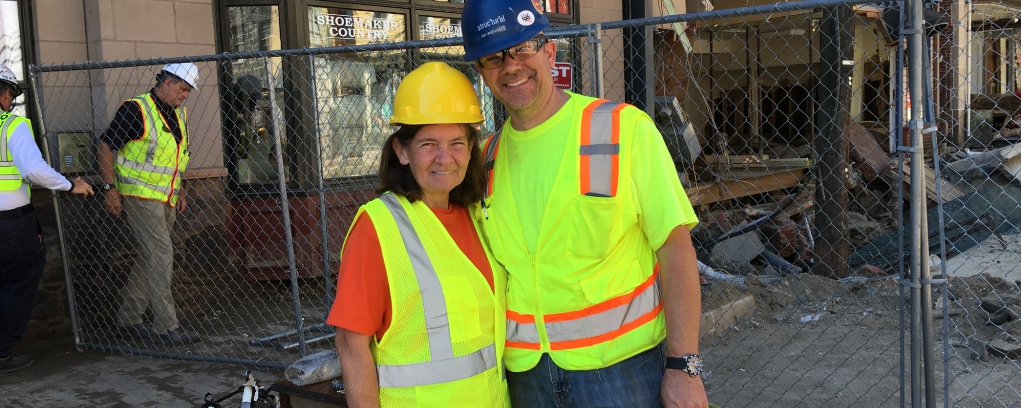 Two people wearing reflective vests and hard hats pose for a photo outside a historic building