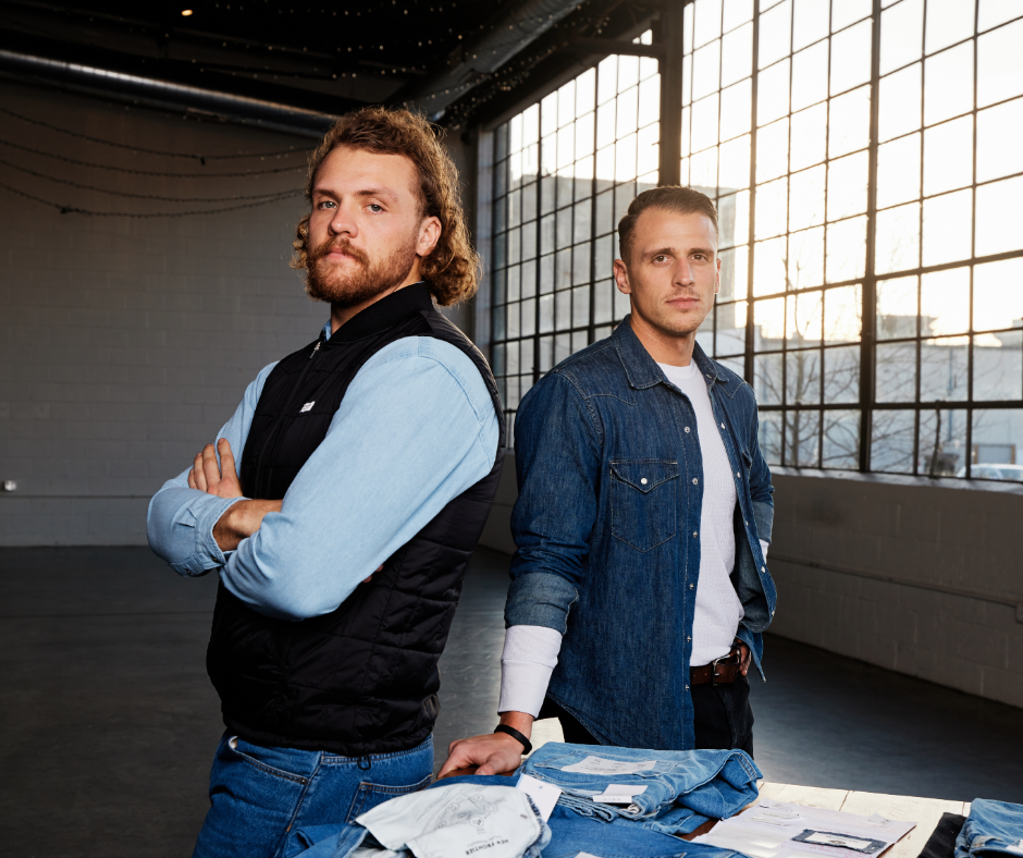 Two men stand back to back behind a table with jeans on it.