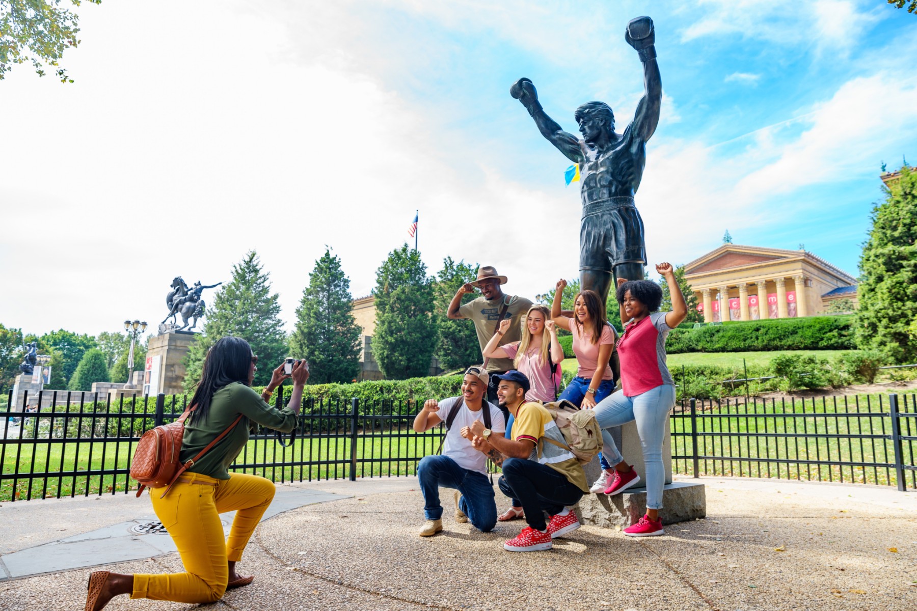 A woman holds up a phone while a group of people pose for a photo in front of the Rocky statue