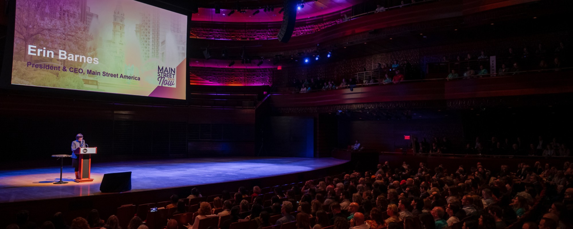 A large crowd sitting in a dimly-lit theater listening to a person speaking from the stage.