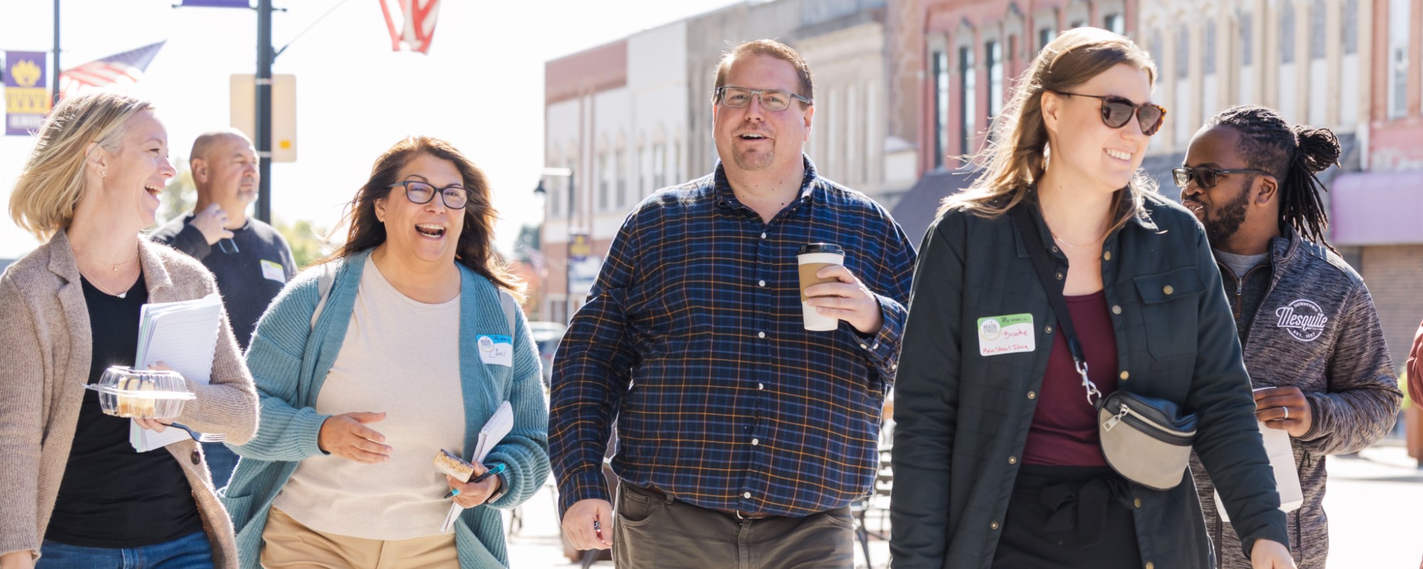 A group of adults laugh while walking through a downtown area with brick buildings and banners visible in the background.
