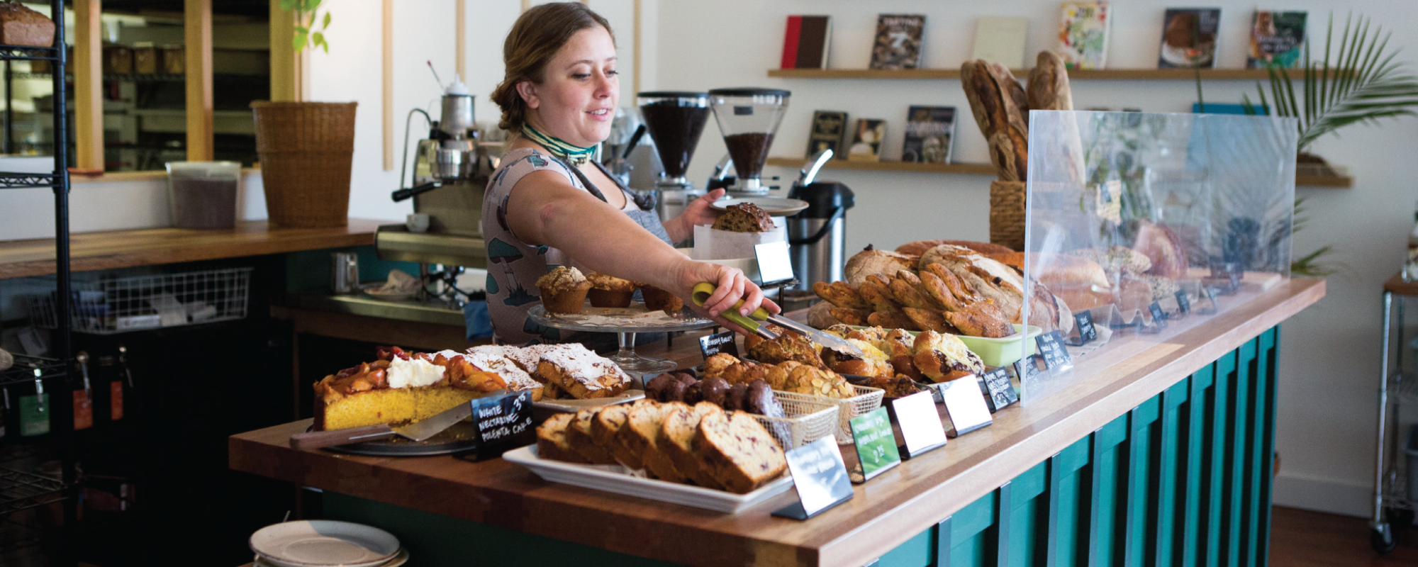 A woman behind a bakery counter uses tongs to pick up bread.