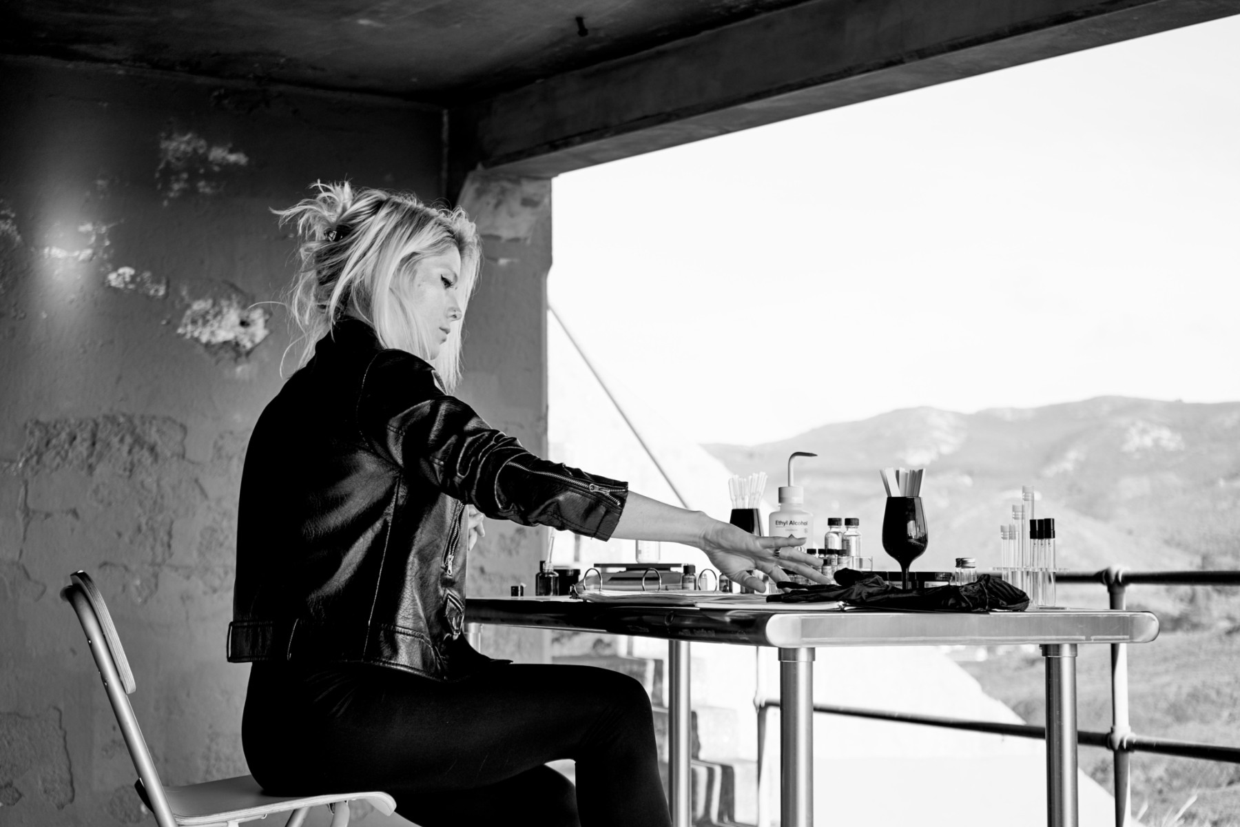 Black and white photo of woman wearing leather jacket sitting at desk with glass bottles.