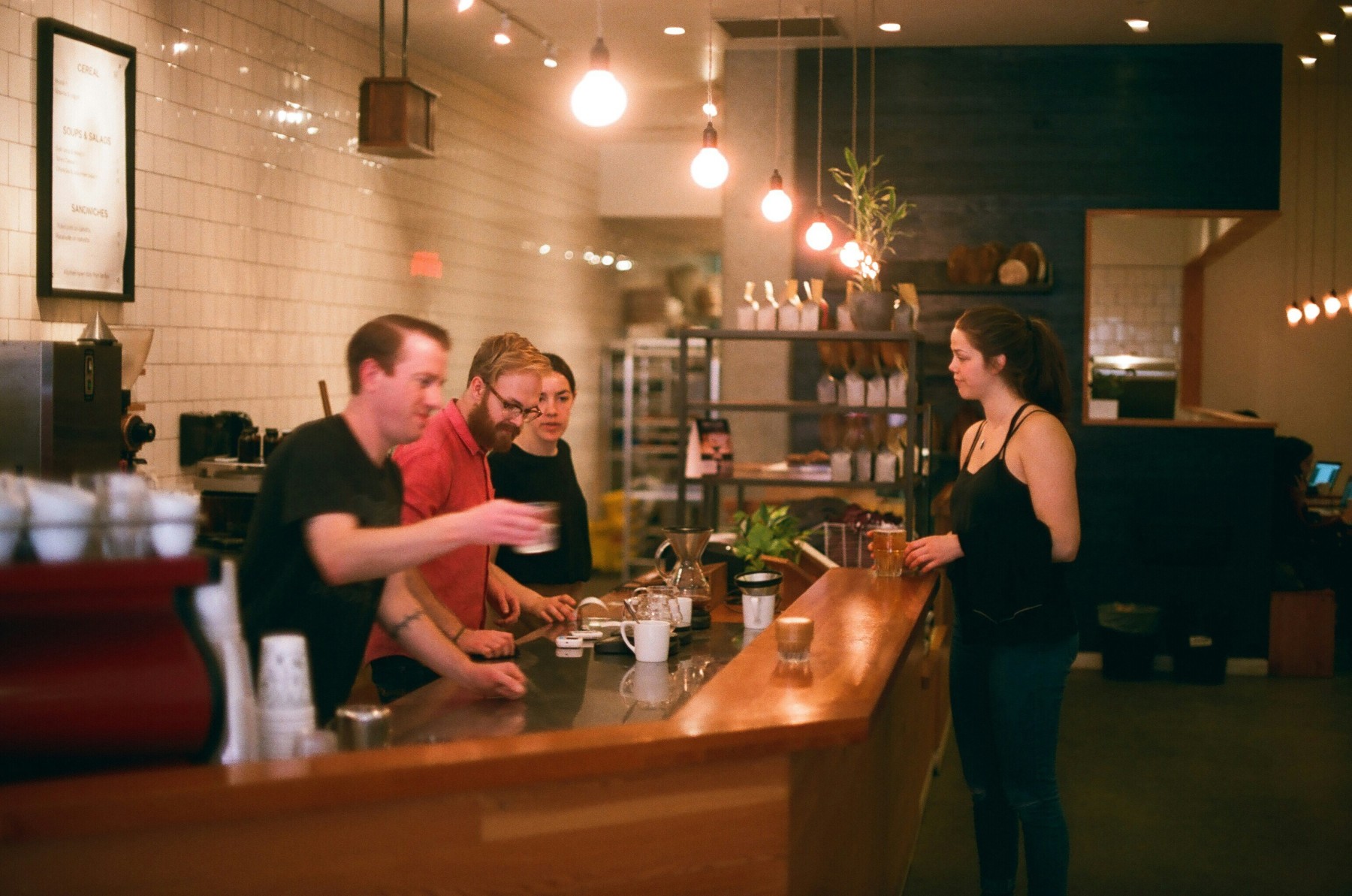 A woman orders coffee in a cafe while men prepare coffee orders behind the counter