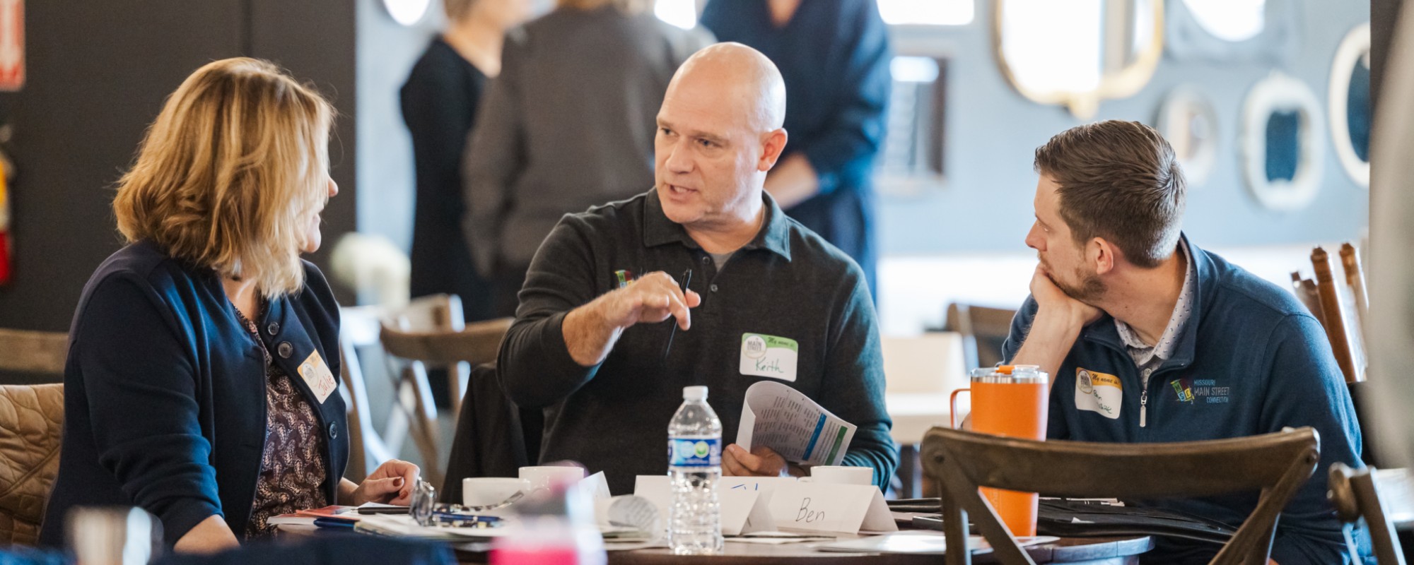 Three people sit around a table; in the center, a man gestures while speaking.