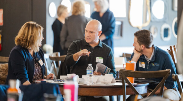 Three people sit around a table; in the center, a man gestures while speaking.