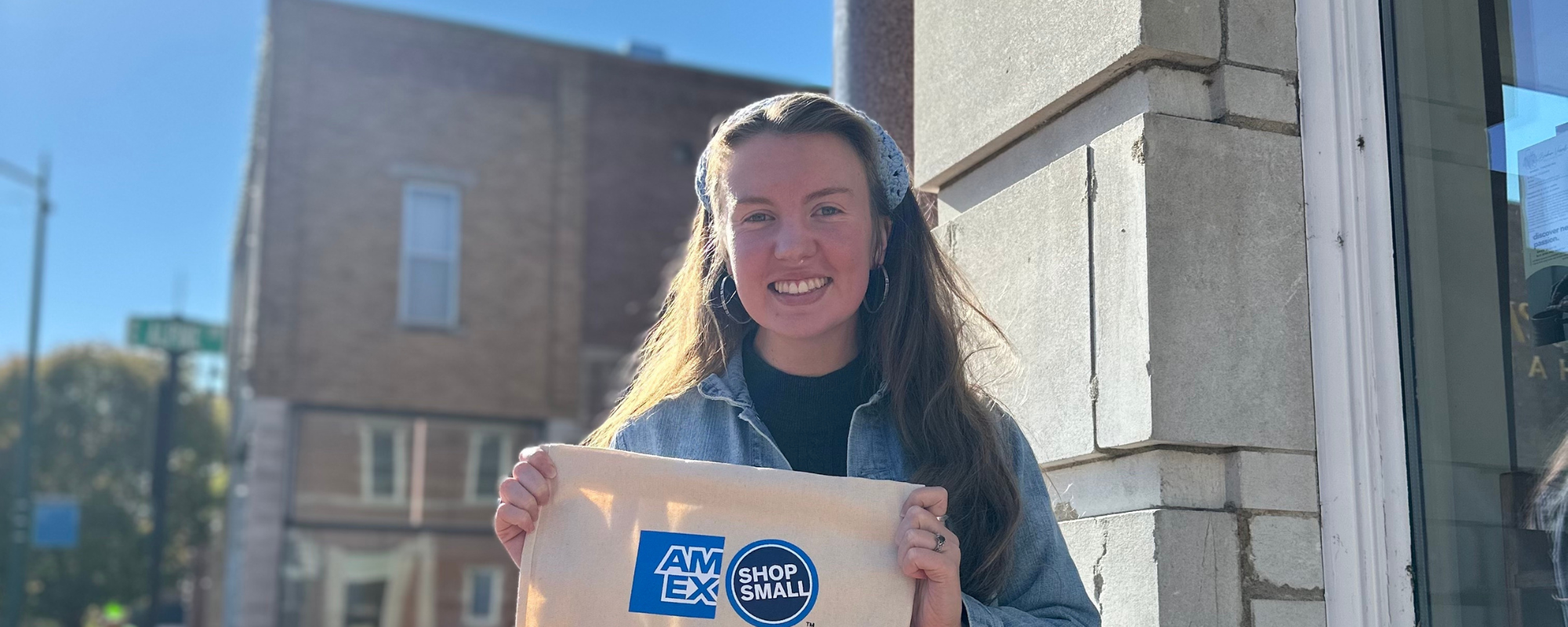 Smiling woman holds bag with "AmEx Shop Small" branding in front of storefront.