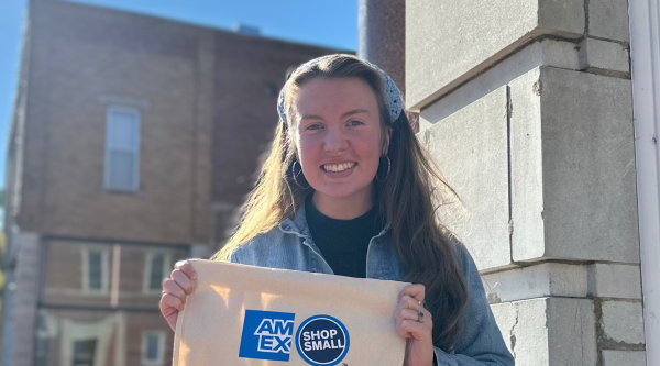 Smiling woman holds bag with "AmEx Shop Small" branding in front of storefront.