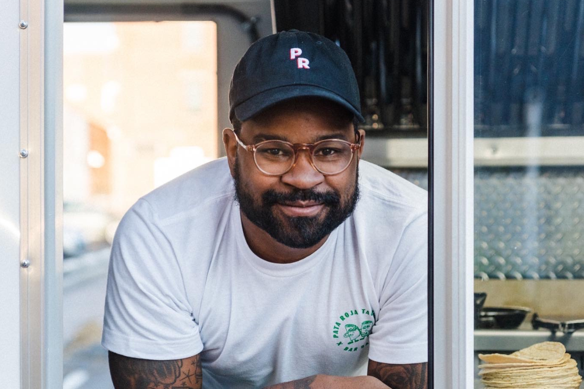 Photo of man wearing blue baseball cap and white t-shirt leaning out the window of a food truck.