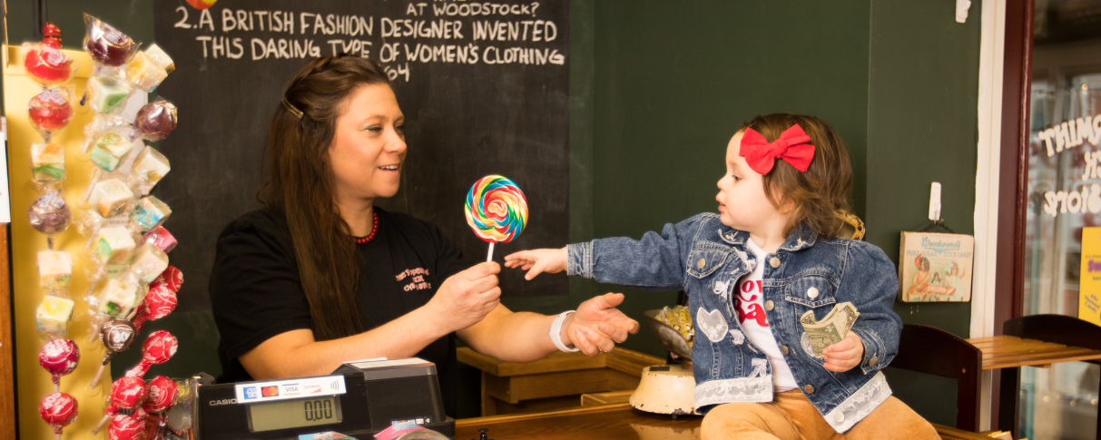 Woman standing behind candy counter hands lollipop to child.