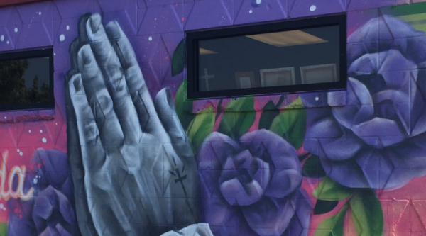 A mural with pair of hands in prayer surrounded by purple roses on a pink and purple background