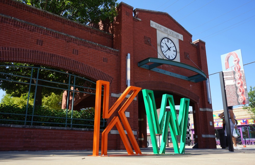 Large metal "K" and "W" letters installed on a sidewalk in front of a brick perimeter wall with windows providing a glimpse into a lush central square.