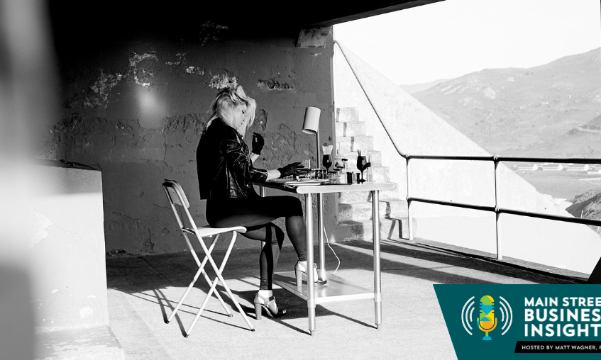 Fotografía en blanco y negro de una mujer sentada ante una pequeña mesa de trabajo. En primer plano hay un gráfico que dice "Main Street Business Insights".