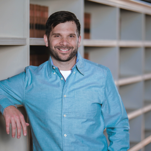 Man in blue shirt leans on bookshelf and smiles at camera