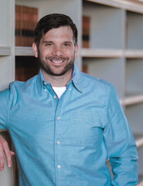 Man in blue shirt leans on bookshelf and smiles at camera